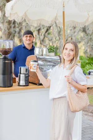 Woman holding a coffee cup at an outdoor coffee cart with barista and espresso machine.