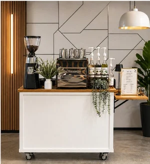 Coffee station with a coffee grinder, glass cups, bottles of flavoring syrups, a menu, and decorative plants in a modern cafe setting.