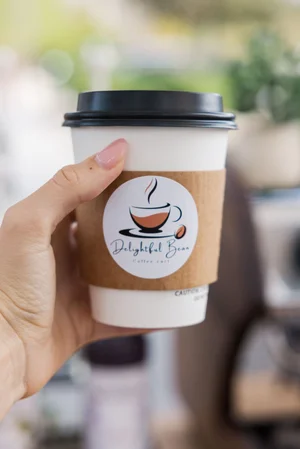Hand holding a coffee cup with "Delightful Bean" logo, black lid, and brown sleeve.