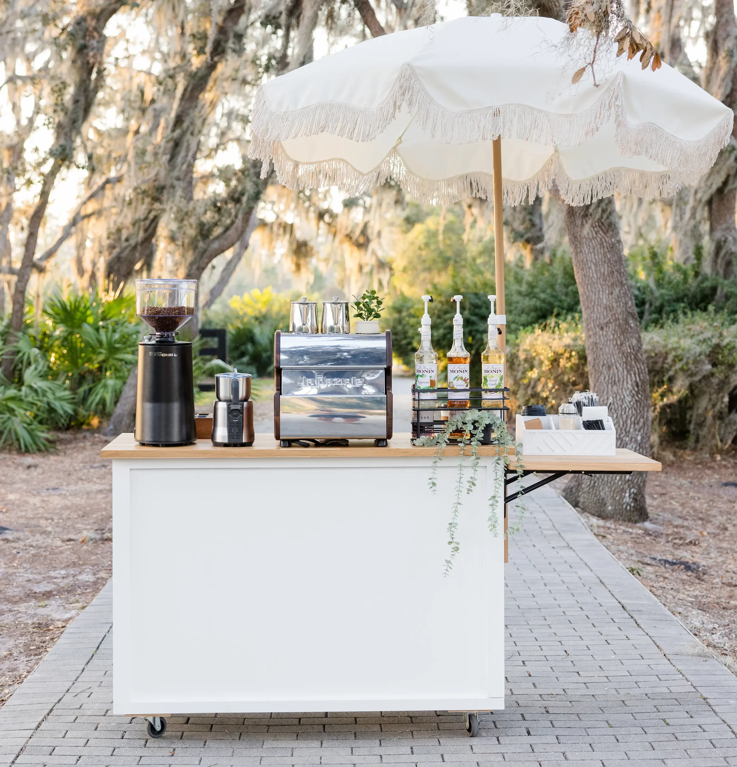 Outdoor beverage station with a white fringed umbrella, a coffee grinder, a food slicer, bottles of flavored syrup, and small serving containers, set on a wooden table on a paved pathway surrounded by trees and greenery.