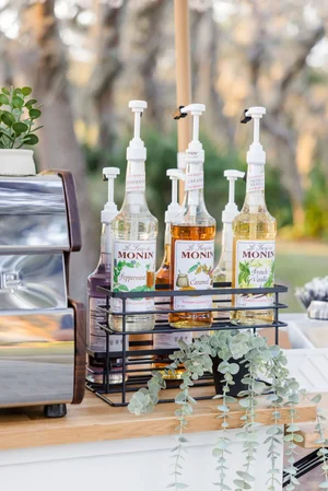 Coffee bar with Monin syrup bottles, peppermint, caramel, and vanilla flavors, alongside coffee machine and decorative plant.
