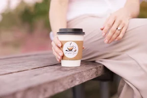 Person holding a coffee cup with "Delightful Bean" logo on a wooden bench.