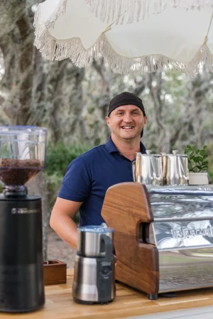 Barista smiling behind a coffee machine outdoors under a fringed umbrella.
