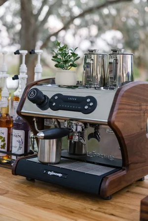Espresso machine with wooden trim, metal milk frothers, a small potted plant, and flavored syrup bottles on a counter.